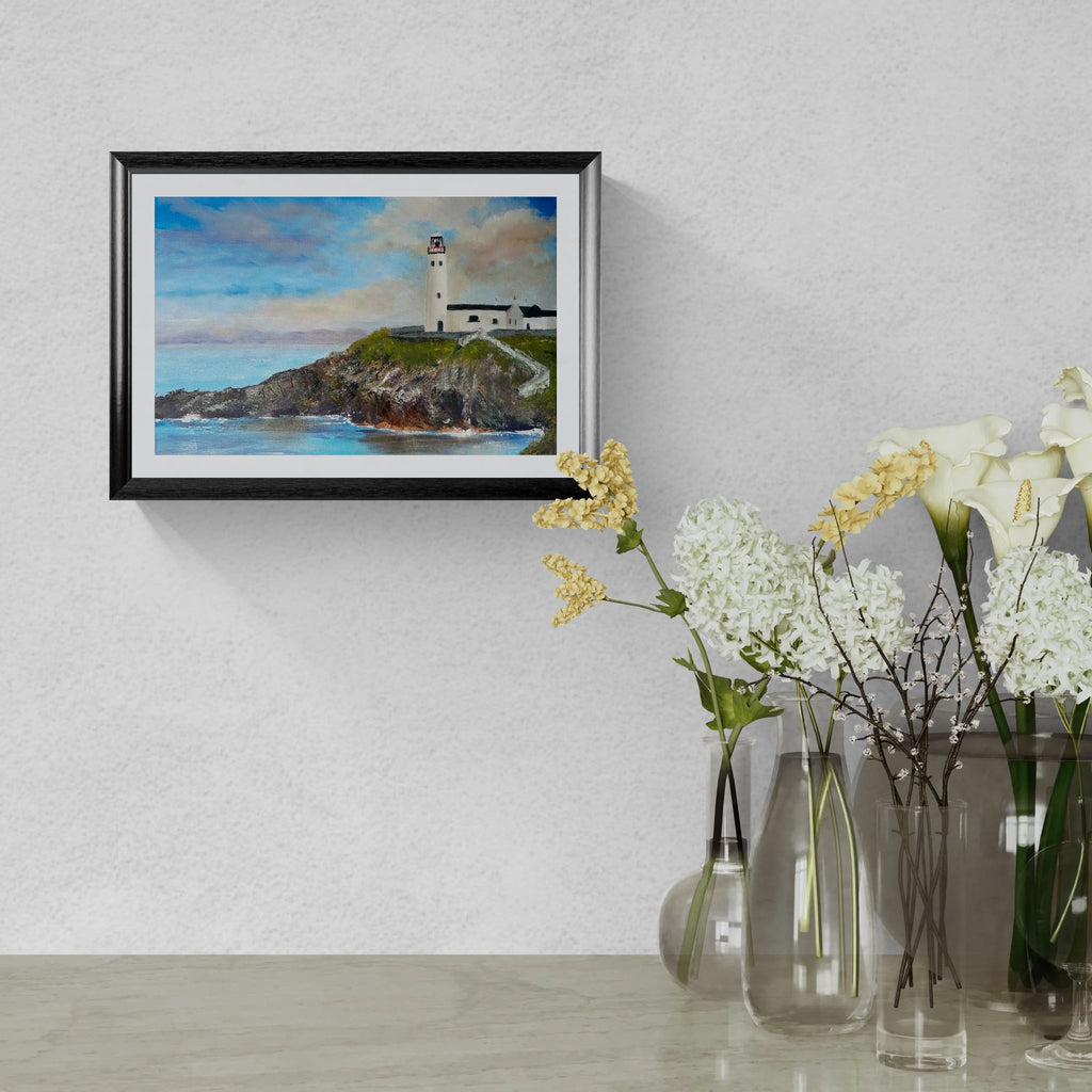 A framed landscape art print of a lighthouse on a rocky cliff under a blue sky, displayed on a textured light gray wall above a table with glass vases and white flowers.