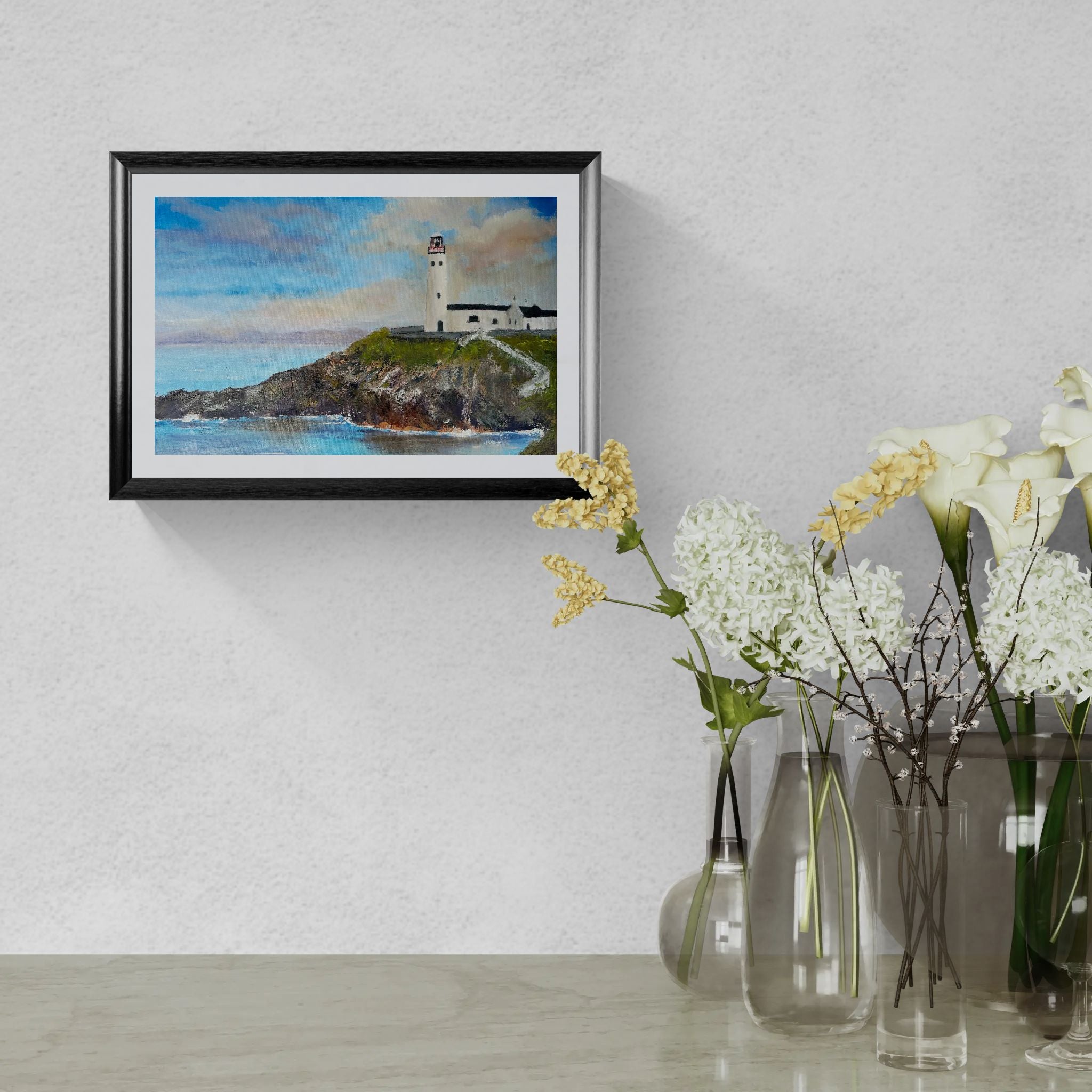 A framed landscape art print of a lighthouse on a rocky cliff under a blue sky, displayed on a textured light gray wall above a table with glass vases and white flowers.