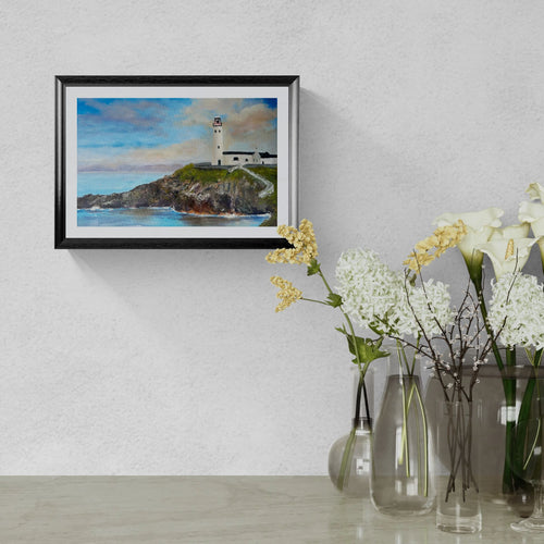 A framed landscape art print of a lighthouse on a rocky cliff under a blue sky, displayed on a textured light gray wall above a table with glass vases and white flowers.