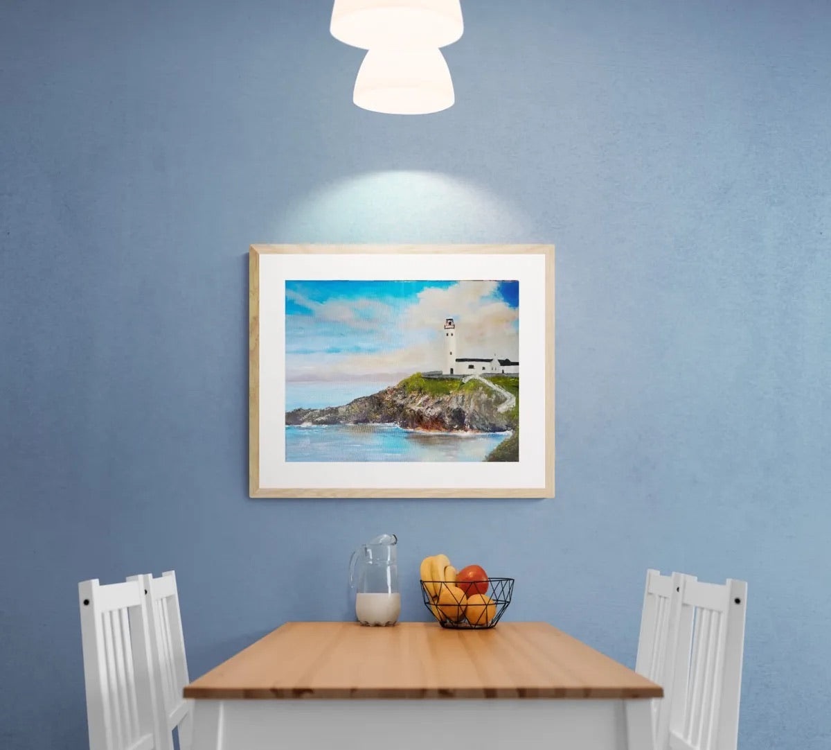 A framed coastal lighthouse painting hangs on a textured blue wall above a wooden dining table with white chairs. The artwork depicts a white lighthouse on rocky green cliffs above blue water under a cloudy sky.