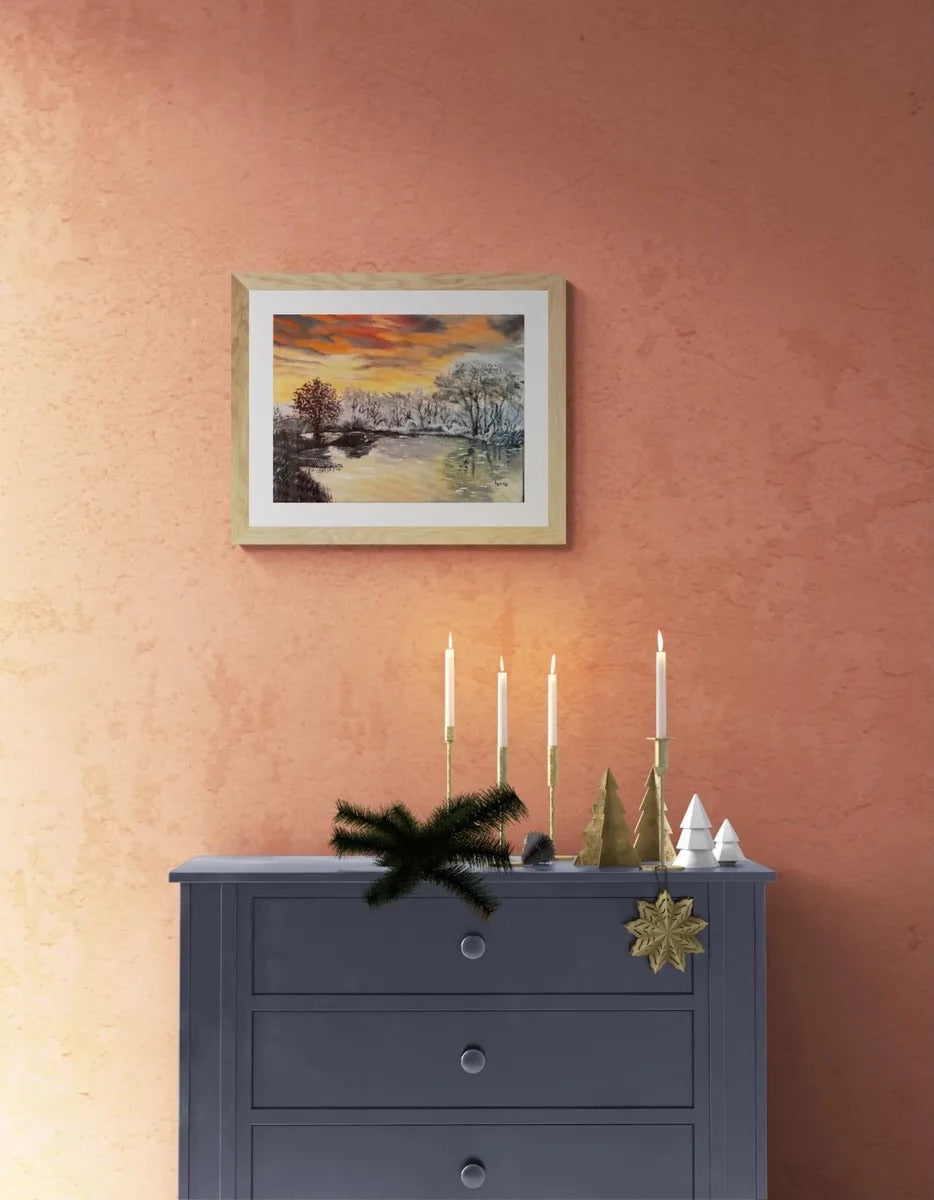 A framed landscape painting of a snowy river at sunset hangs on a textured terracotta wall above a dark gray dresser decorated with candles, miniature trees, and holiday ornaments.