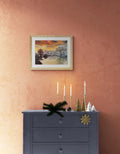 A framed landscape painting of a snowy river at sunset hangs on a textured terracotta wall above a dark gray dresser decorated with candles, miniature trees, and holiday ornaments.